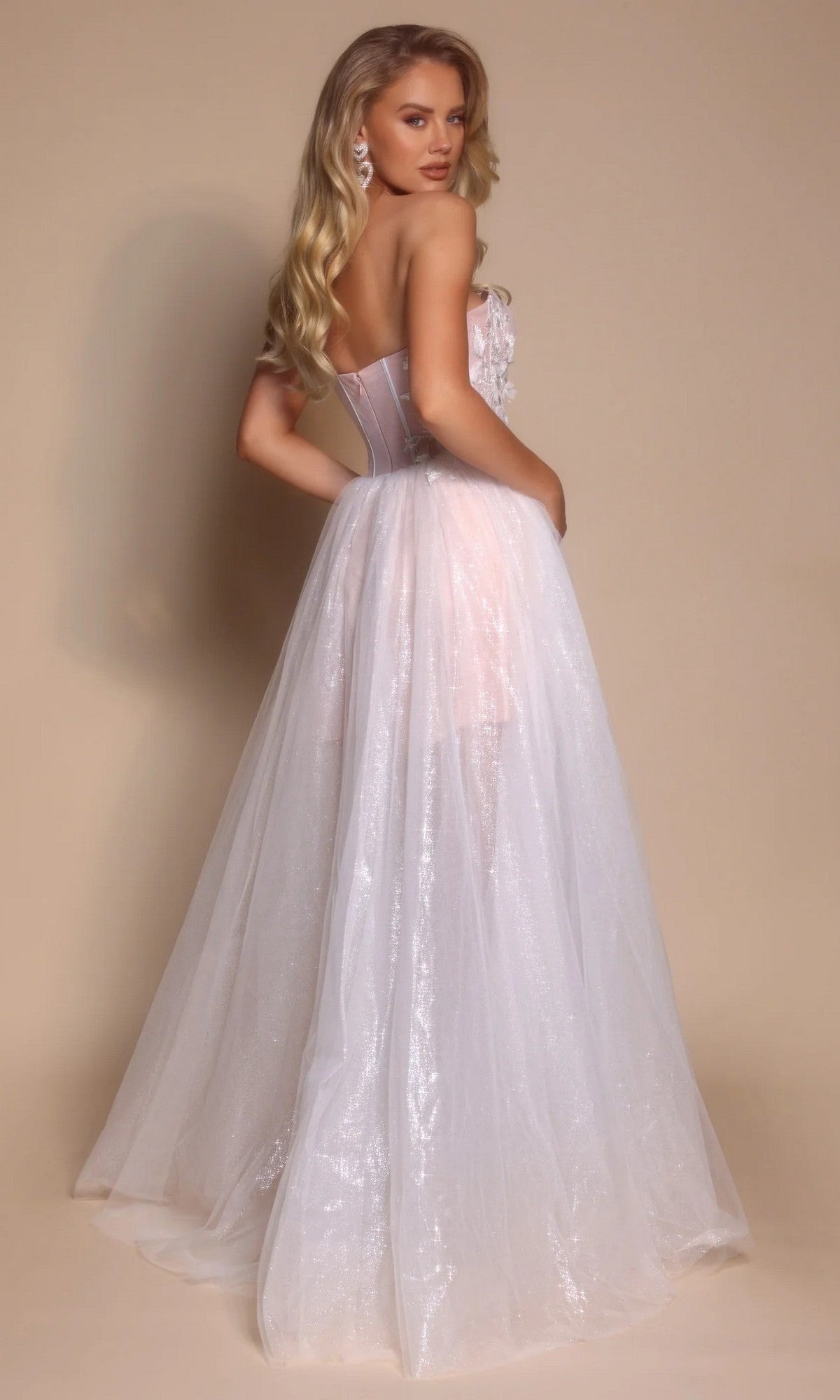 A woman with long blonde hair poses against a plain beige background wearing the Long Formal Dress: Portia and Scarlett PS26078, an ethereal gown featuring a corset bodice and sheer white fabric with floral details.